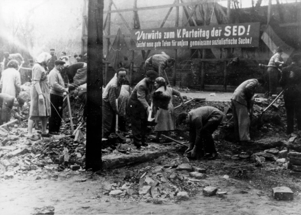 Umbauarbeiten im Prater 1958. Viele freiwilligen Helfer:innen des Nationalen Aufbauwerks arbeiten mit Werkzeug in den Trümmern. Im Hintergrund hängt ein Werbespruch der SED.