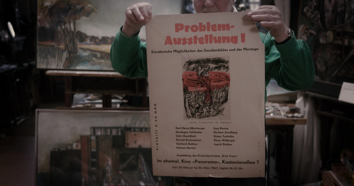 Skip Pahler hält in seinem Atelier das Plakat zur ersten Ausstellung der Galerie von 1967 in die Kamera. 